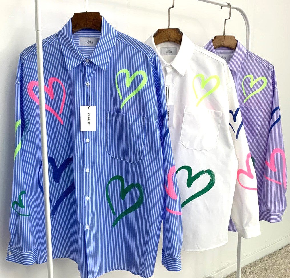 Blue, white, purple collared shirts with heart graphics and front pockets hanging on a rack. 